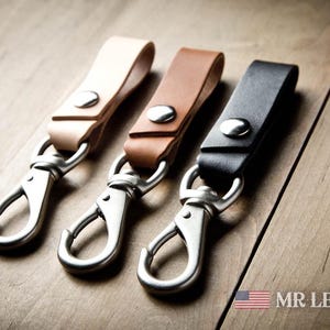 Pode incluir: Tr&ecirc;s porta-chaves de couro em bege claro, castanho m&eacute;dio e preto, cada um com um bot&atilde;o de press&atilde;o de metal prateado e um mosquet&atilde;o girat&oacute;rio. O texto MR LENTZ.com e um pequeno &iacute;cone da bandeira americana est&atilde;o vis&iacute;veis no canto inferior.