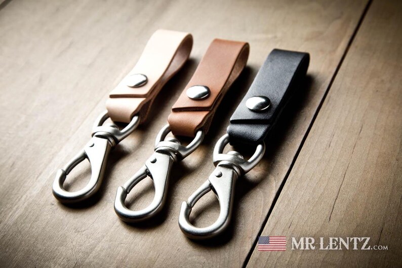 Pode incluir: Tr&ecirc;s porta-chaves de couro em bege claro, castanho m&eacute;dio e preto, cada um com um bot&atilde;o de press&atilde;o de metal prateado e um mosquet&atilde;o girat&oacute;rio. O texto MR LENTZ.com e um pequeno &iacute;cone da bandeira americana est&atilde;o vis&iacute;veis no canto inferior.
