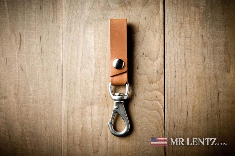 Pode incluir: Uma al&ccedil;a de chaveiro em couro bege com um bot&atilde;o de press&atilde;o de metal prateado e um mosquet&atilde;o girat&oacute;rio. A al&ccedil;a &eacute; dobrada e fixada com as ferragens met&aacute;licas, proporcionando um ponto de fixa&ccedil;&atilde;o funcional para chaves ou outros pequenos itens.