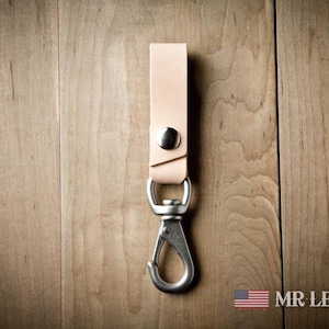Pode incluir: Um porta-chaves de couro castanho claro com um bot&atilde;o de press&atilde;o de metal prateado e um mosquet&atilde;o girat&oacute;rio. A tira de couro est&aacute; dobrada e fixada com as ferragens met&aacute;licas. O texto MR LENTZ.COM est&aacute; impresso no canto inferior direito.