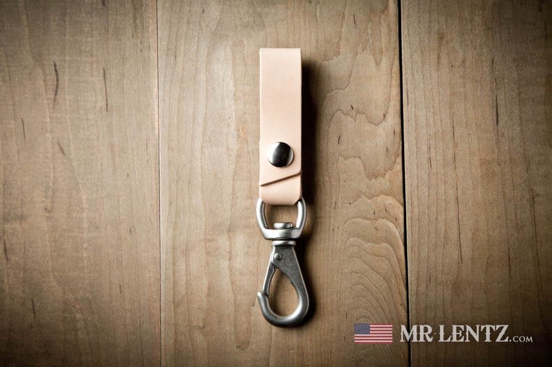 Pode incluir: Um porta-chaves de couro castanho claro com um bot&atilde;o de press&atilde;o de metal prateado e um mosquet&atilde;o girat&oacute;rio. A tira de couro est&aacute; dobrada e fixada com as ferragens met&aacute;licas. O texto MR LENTZ.COM est&aacute; impresso no canto inferior direito.