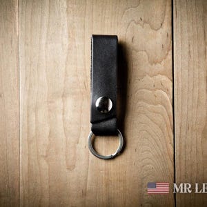 Pode incluir: Uma tira de porta-chaves em couro preto com um fecho de bot&atilde;o de press&atilde;o em metal prateado e um anel circular em metal prateado na parte inferior. O texto MRLENTZ.COM &eacute; vis&iacute;vel no canto inferior direito junto a uma bandeira dos EUA.