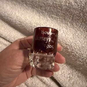 May include: A small, vintage shot glass with a clear base and a dark red upper portion. The glass has etched text that reads "Souvenir Jerseyville 1905". The glass is held in a hand against a white, textured background.