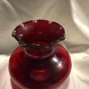 以下が含まれることがあります： ルビーレッドのガラスの花瓶、フリル付きの縁。花瓶は丸いボディとフレアの入ったスカラップトップを備えています。ガラスは滑らかで光沢があり、光を反射しているように見えます。花瓶はおそらく装飾品です。