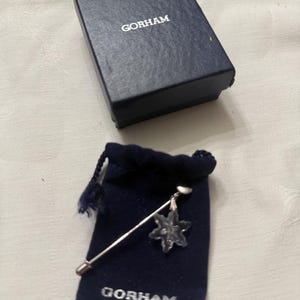 May include: A silver-toned pin with a clear, snowflake-shaped crystal charm, displayed with a navy blue velvet pouch and a navy blue box. The box and pouch are both labeled "GORHAM" in white lettering. The pin is a decorative accessory.