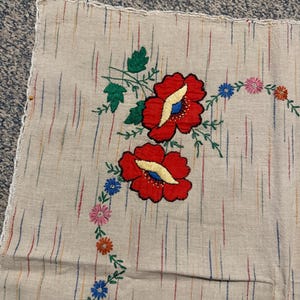 Pode incluir: Um quadrado de tecido bege com linhas verticais tracejadas multicoloridas e motivos florais bordados. Duas grandes papoulas vermelhas com centros amarelos e folhas verdes estão centralizadas, rodeadas por uma trepadeira de pequenas flores rosas, azuis e laranjas.