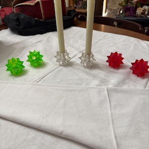 May include: A table setting featuring two ivory-colored candles in clear star-shaped holders. Two green star-shaped objects and three red star-shaped objects are also on the white tablecloth. The candles have a flickering flame effect.