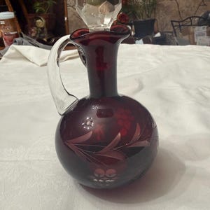 May include: A ruby red glass decanter with a clear glass handle and stopper. The decanter has etched floral designs and a faceted stopper. The handle is curved, and the decanter has a round body. The stopper is a large, clear, multi-faceted crystal.
