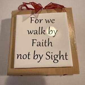 May include: A decorative square tile with the text "For we walk by Faith not by Sight" in black lettering. The tile is set on a wooden frame and adorned with a red ribbon.