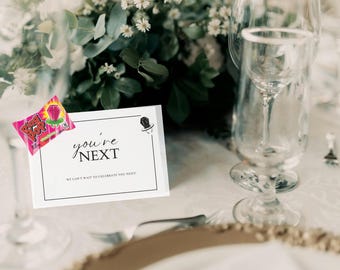 May include: A white rectangular card with a black border featuring the text you're NEXT and WE CAN'T WAIT TO CELEBRATE YOU NEXT! with a small top hat icon, accompanied by a pink strawberry Ring Pop candy wrapper positioned on the left corner.
