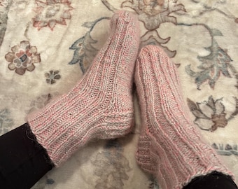 May include: A pair of pink and gray knitted socks. The socks have a ribbed texture and are worn on a patterned rug. The socks are a light pink color with gray accents, and appear to be handmade.