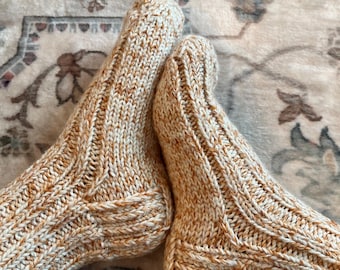 May include: A pair of hand-knitted socks in a warm, speckled color palette of cream and light brown. The socks feature a ribbed design on the leg and foot, with a reinforced heel and toe. The cozy socks are perfect for cold weather.