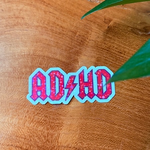 May include: A sticker with the word "ADHD" in a red, stylized font with a lightning bolt, on a light blue background. The sticker has a holographic effect and is placed on a wooden surface.