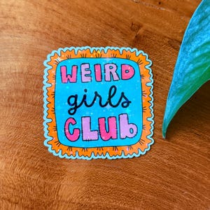 May include: A sticker with the text "WEIRD girls CLUB" in pink and blue. The text is on a blue background with a glitter effect, framed by an orange border with a wavy edge. The sticker is on a wooden surface.