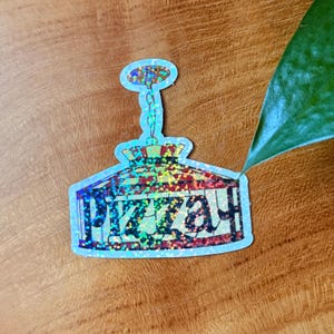 Op de afbeelding: Holografische sticker met een gestileerd pizza-ovenontwerp met het woord "PIZZAH" in een speels lettertype. De sticker heeft een lichtblauwe rand en een kleurrijk, glinsterend interieur. De sticker ligt op een houten oppervlak.