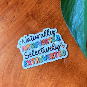 May include: A light blue sticker with the words "Naturally Introverted, Selectively Extroverted" in a colorful, holographic font. The sticker has a star and diamond design.