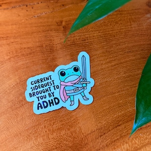 May include: A sticker featuring a cartoon frog warrior with a sword, wearing a pink cape and scarf. The sticker has the text "CURRENT SIDEQUEST BROUGHT TO YOU BY ADHD" in blue and white.