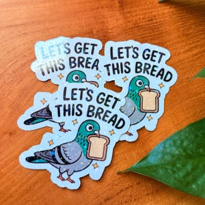 May include: A collection of holographic stickers featuring a cartoon pigeon holding a slice of bread. The text "LET'S GET THIS BREAD" is printed above the pigeon. The stickers have a blue, green, and gray color scheme.