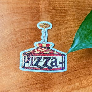 Op de afbeelding: Een sticker in de vorm van een pizzalamp. De lamp heeft een rood en geel geblokt patroon, met het woord "PIZZA" in een glitterlettertype. De sticker heeft een lichtblauwe rand.