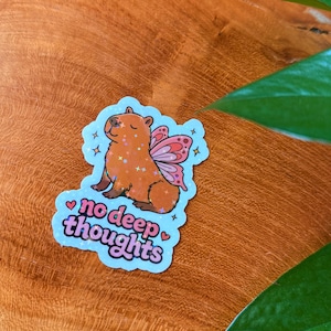 May include: A sticker featuring a cartoon capybara with fairy wings, glitter accents, and the text "no deep thoughts" in a playful font. The sticker has a light blue border with star and heart details.