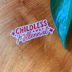 May include: A white sticker with the words "CHILDLESS Millennial" in red, glittery lettering. The sticker has a white border and is decorated with small hearts, stars, and diamond shapes. The sticker is on a wooden surface.