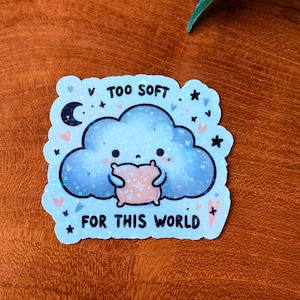 May include: A light blue sticker with a cloud design holding a pink pillow. The sticker has the text "TOO SOFT FOR THIS WORLD" and is decorated with stars, hearts, and a crescent moon.