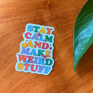 May include: A colorful sticker with the text "STAY CALM AND MAKE WEIRD STUFF" in a retro font. The sticker has a light blue background and features various colors like red, yellow, and purple. A green leaf is visible in the background.
