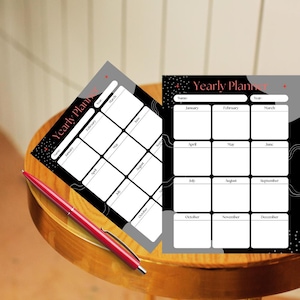 May include: Two black and white yearly planner sheets with the text "Yearly Plan" and a red pen on a wooden table. The planners have a grid layout with months listed. The pen is red.