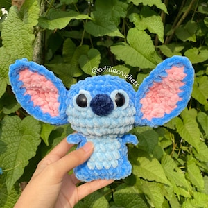 Puede incluir: Mu&ntilde;eco Stitch tejido a ganchillo en tonos azules y rosas, con grandes orejas y ojos negros. El mu&ntilde;eco se sostiene frente a un fondo de hojas verdes. El texto "@odiliacrochets" es visible.