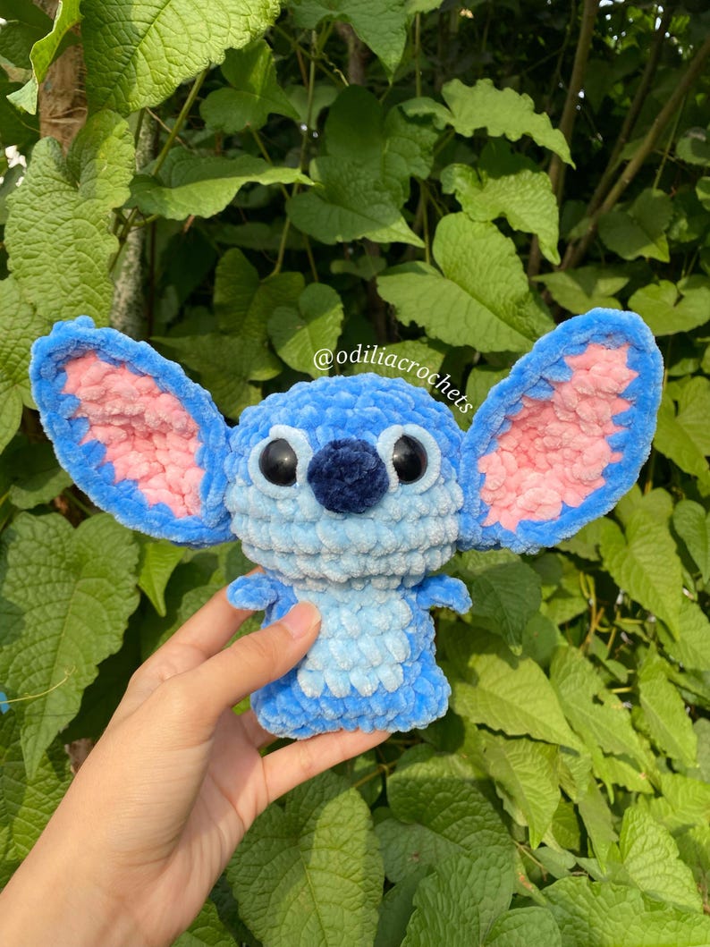 Puede incluir: Mu&ntilde;eco Stitch tejido a ganchillo en tonos azules y rosas, con grandes orejas y ojos negros. El mu&ntilde;eco se sostiene frente a un fondo de hojas verdes. El texto "@odiliacrochets" es visible.