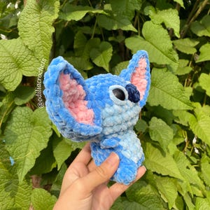 Puede incluir: Un personaje tejido a ganchillo azul y rosa con orejas grandes y ojos negros. El peluche se sostiene contra un fondo de follaje verde. El texto "@odiliacrochets" es visible en el lado izquierdo de la imagen.