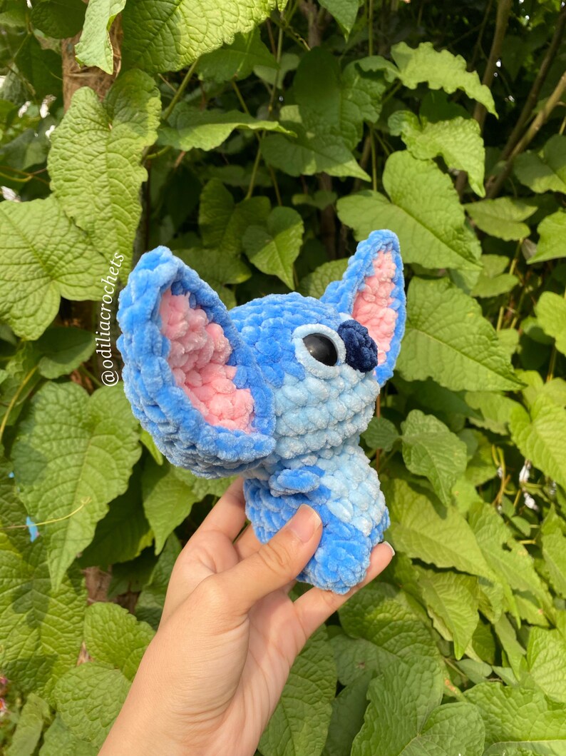 Puede incluir: Un personaje tejido a ganchillo azul y rosa con orejas grandes y ojos negros. El peluche se sostiene contra un fondo de follaje verde. El texto "@odiliacrochets" es visible en el lado izquierdo de la imagen.