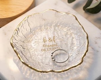 May include: A clear, textured glass trinket dish with a gold-painted rim. The center features gold text reading b & f, Engaged, and 08.17.2024. A simple silver-toned metal ring rests inside the dish, which has an irregular, organic circular shape.