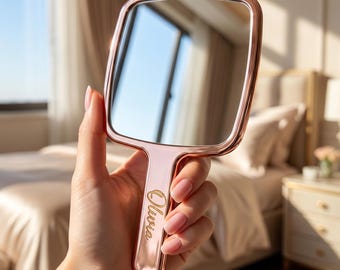 May include: A rose gold hand mirror with a rectangular mirror and a handle. The handle is engraved with the name "Olivia" in a delicate script. The mirror reflects a bright window and a bedroom setting.