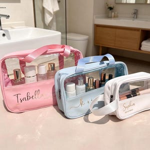 Op de afbeelding: Drie cosmetische tassen in roze, lichtblauw en wit, elk met een doorzichtig voorpaneel en een handvat. De roze tas is gelabeld met "Isabella", de lichtblauwe tas heeft "k + i", en de witte tas zegt "Sophia". Elke tas bevat cosmetische producten.