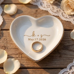 May include: A heart-shaped, white ceramic ring dish with gold trim. The dish has the engraved initials "k + s" and the date "May.17.2026". A gold wedding ring rests inside the dish. The background includes white rose petals and lace.