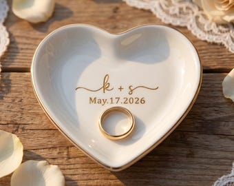 May include: A heart-shaped, white ceramic ring dish with gold trim. The dish has the engraved initials "k + s" and the date "May.17.2026". A gold wedding ring rests inside the dish. The background includes white rose petals and lace.