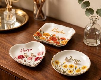 May include: Three white ceramic trinket dishes with gold-toned rims. One heart-shaped dish features red flowers and the name Harper. A square dish displays orange flowers and the name Kaylee. A round dish shows yellow flowers and the name Jessie.