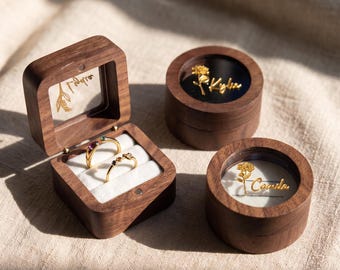 May include: Three dark wood ring boxes with clear lids. One square box holds two gold rings with small stones on a white velvet insert. Two round boxes feature gold floral engravings and the names Kyla and Camila on the clear lids.