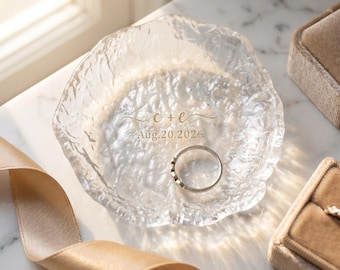 May include: A clear, textured glass ring dish featuring the gold-printed text c + e and Aug.20.2026. A silver-toned metal ring rests inside the dish, which has an irregular, organic edge and a rippled surface that catches the light.