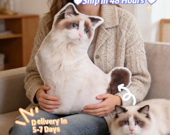 May include: A plush cat-shaped pillow with a white and brown pattern, held by a person. The pillow resembles a Ragdoll cat, with blue eyes and a fluffy tail. A real Ragdoll cat is also in the image. The text "Ship in 48 Hours" is visible.