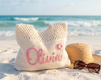 May include: A cream-colored crocheted beach bag with the name "Olivia" embroidered in pink. A small pink flower is attached to the bag. A straw hat and tortoiseshell sunglasses rest on the sand next to the bag, with the ocean in the background.