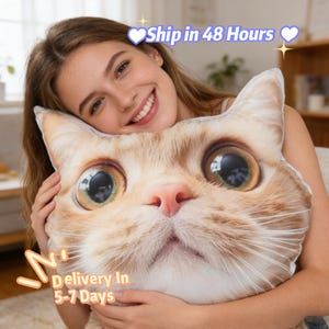 Peut inclure: Un coussin en forme de chat avec une impression réaliste d'un visage de chat, avec de grands yeux et un nez rose. Le coussin est tenu par une personne. Le texte "Ship in 48 Hours" et "Delivery In 5-7 Days" sont également visibles.