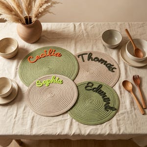 May include: Four round, coiled rope placemats in sage green and beige. Each placemat features a personalized name in raised, cursive yarn lettering: Cecilia in orange, Thomas in dark brown, Sophia in lime green, and Edmund in dark brown.