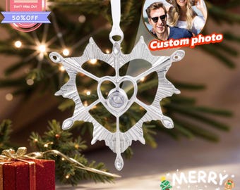 May include: A silver snowflake ornament with a heart-shaped center and a clear gem. The ornament hangs from a white ribbon. The image also includes a red gift box, a "Merry Christmas" graphic, and a "Custom photo" option.