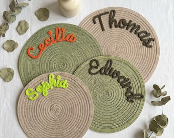 May include: Four circular woven rope placemats in olive green and beige. Each placemat features a personalized name in raised, cursive yarn lettering: Cecilia in orange, Thomas in dark brown, Sophia in lime green, and Edward in dark brown.