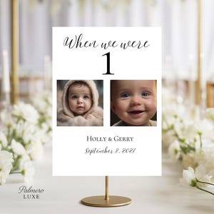 May include: A white table sign with the text "When we were 1" above two baby photos. The sign includes the names "Holly & Gerry" and the date "September 8, 2027". The sign is on a gold stand.