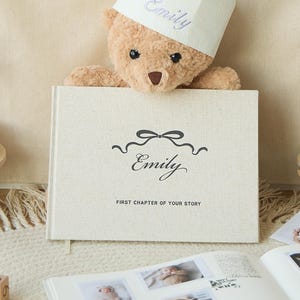 Peut inclure: Scène d'anniversaire avec un ours en peluche portant une couronne avec "Emily". Un livre avec "Emily" sur la couverture est devant l'ours. L'arrière-plan indique "Happy Birthday Emily". Un présentoir à gâteau en bois et un album photo sont également présents.
