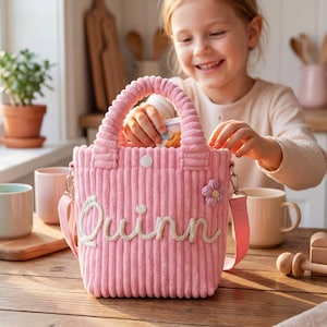 May include: A pink corduroy tote bag with the name "Quinn" embroidered in white script. The bag features a top handle, a shoulder strap, and a flower applique. A young girl is smiling and holding the bag.