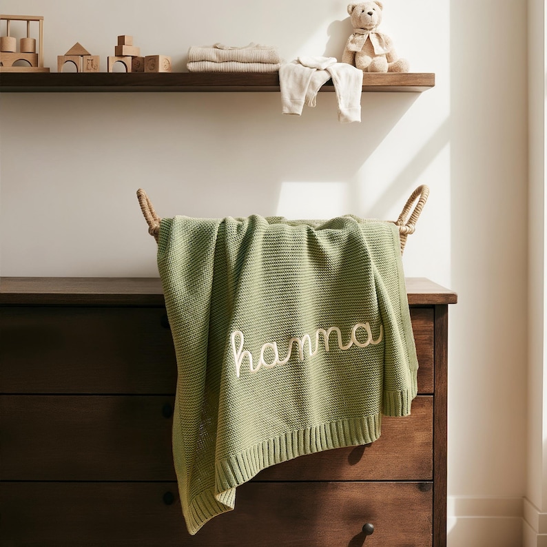 May include: A sage green knitted baby blanket embroidered with the name "hanna" in white. The blanket is draped over a wicker basket on a wooden chest of drawers. A shelf above displays baby clothes and toys.
